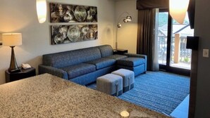A modern hotel room with a large sofa, a small ottoman, a granite countertop, a ceiling fan, and a large window with a view of the outside.