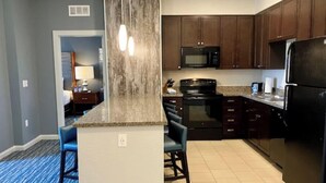 A modern kitchen with dark wooden cabinets, a black refrigerator, a microwave, and a stove. There is a blue chair and a small island with a light-colored countertop.