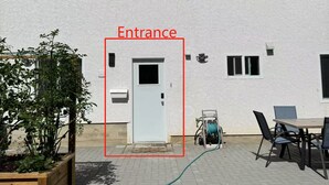 Un edificio con fachada blanca, entrada señalizada, patio con mesa y sillas, y jardín con plantas.