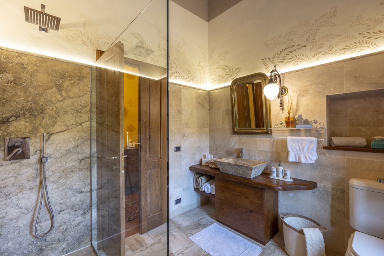 A bathroom with a glass shower enclosure, a wooden vanity, a marble-tiled floor, and a wall-mounted mirror.