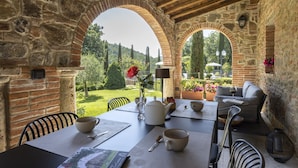 Un patio al aire libre con paredes de piedra, una mesa dispuesta con un hervidor, tazas y un jarrón con flores.