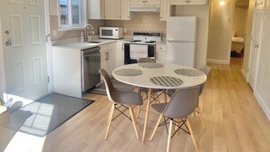 Une cuisine compacte avec des meubles blancs, un four à micro-ondes, une cuisinière et un réfrigérateur. On y trouve une table à manger avec quatre chaises et une petite fenêtre.