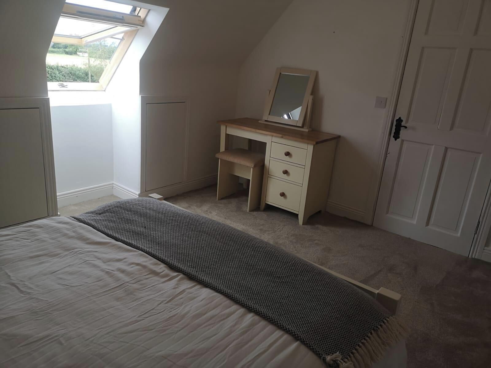 A bedroom with a bed, a desk with a mirror, a door, and a window.