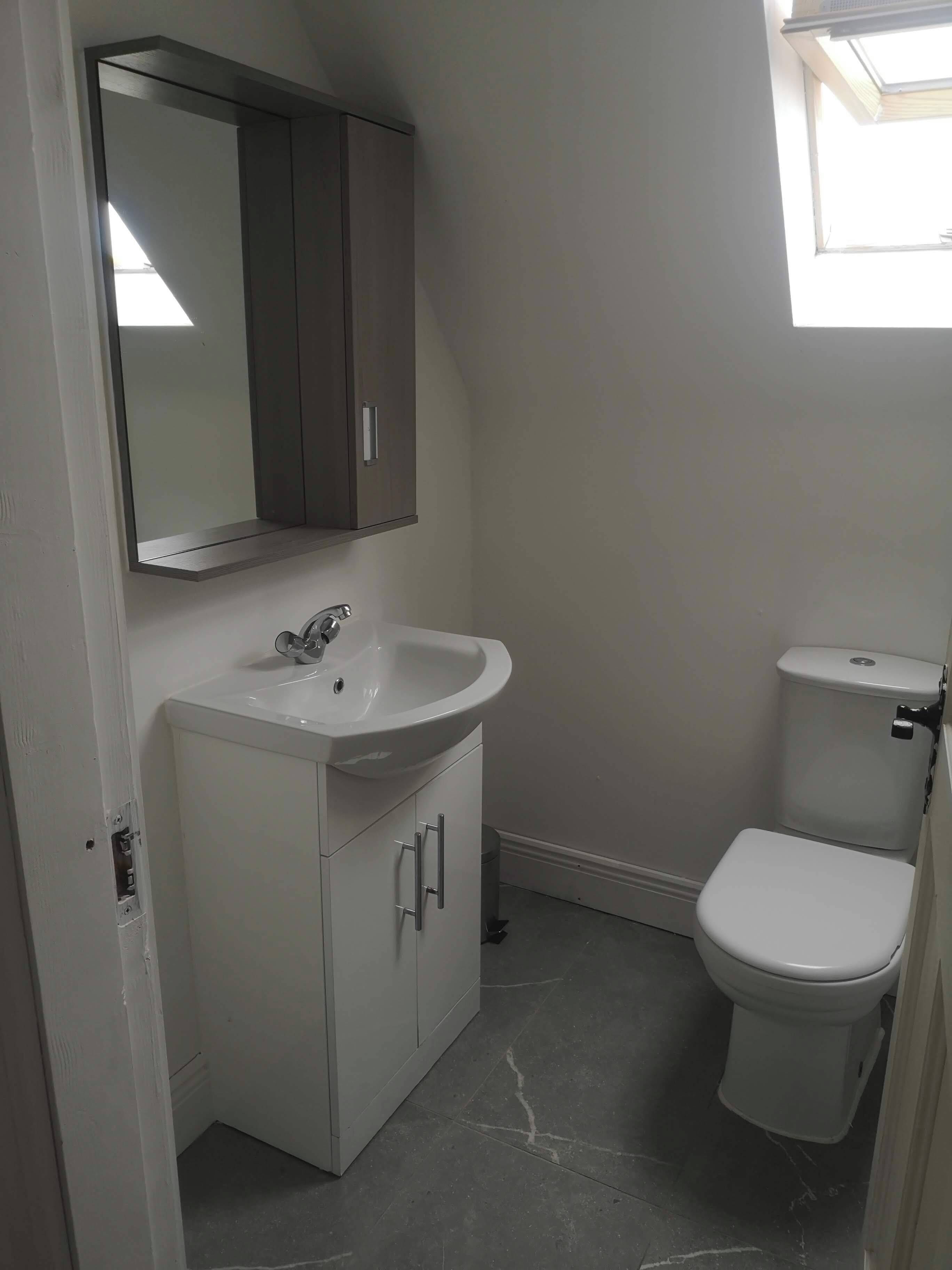 A bathroom with a white sink, a mirror above it, a toilet, and a window providing natural light.