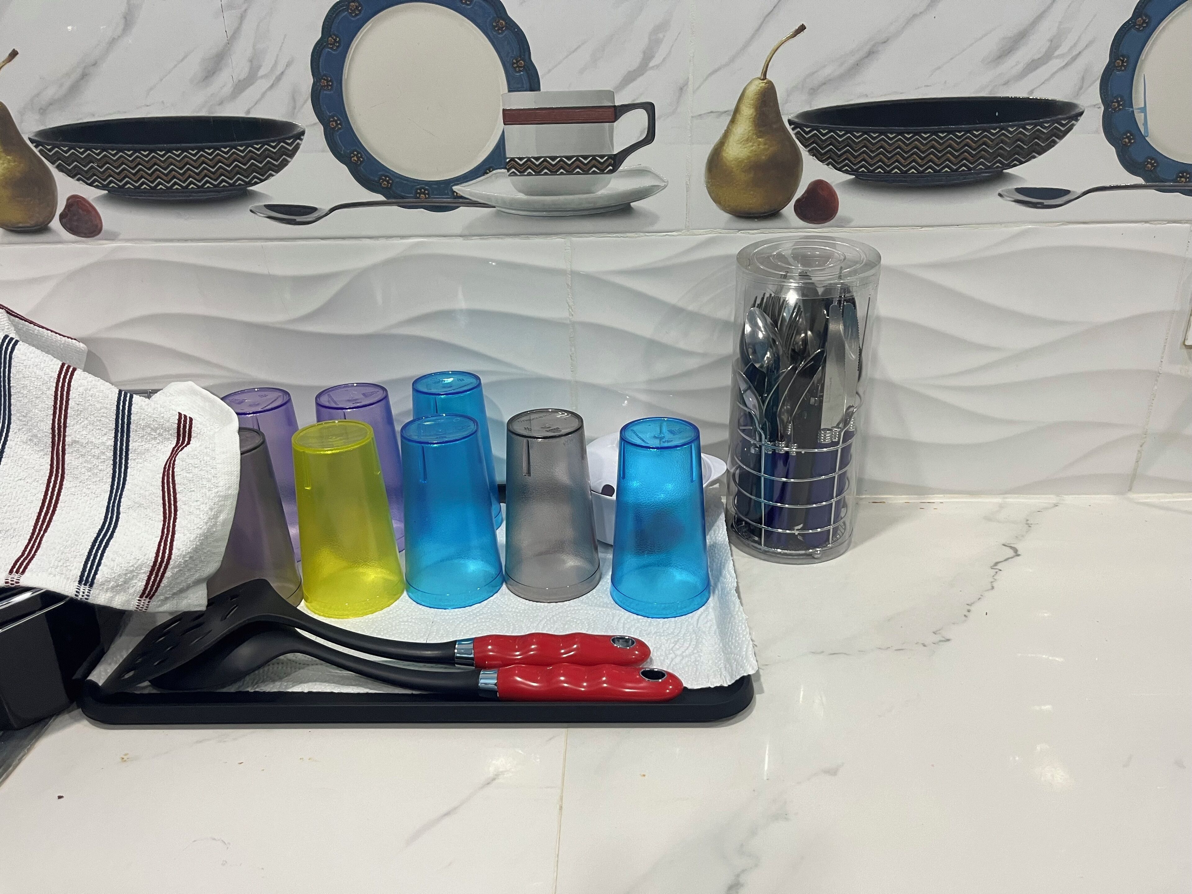 Een aanrecht met verschillende glazen bekers in diverse kleuren, een metalen houder met keukengerei en een gestreepte handdoek.