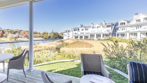 Un espacio al aire libre cubierto con muebles de mimbre, vista a un lago y una fila de casas grandes al fondo.
