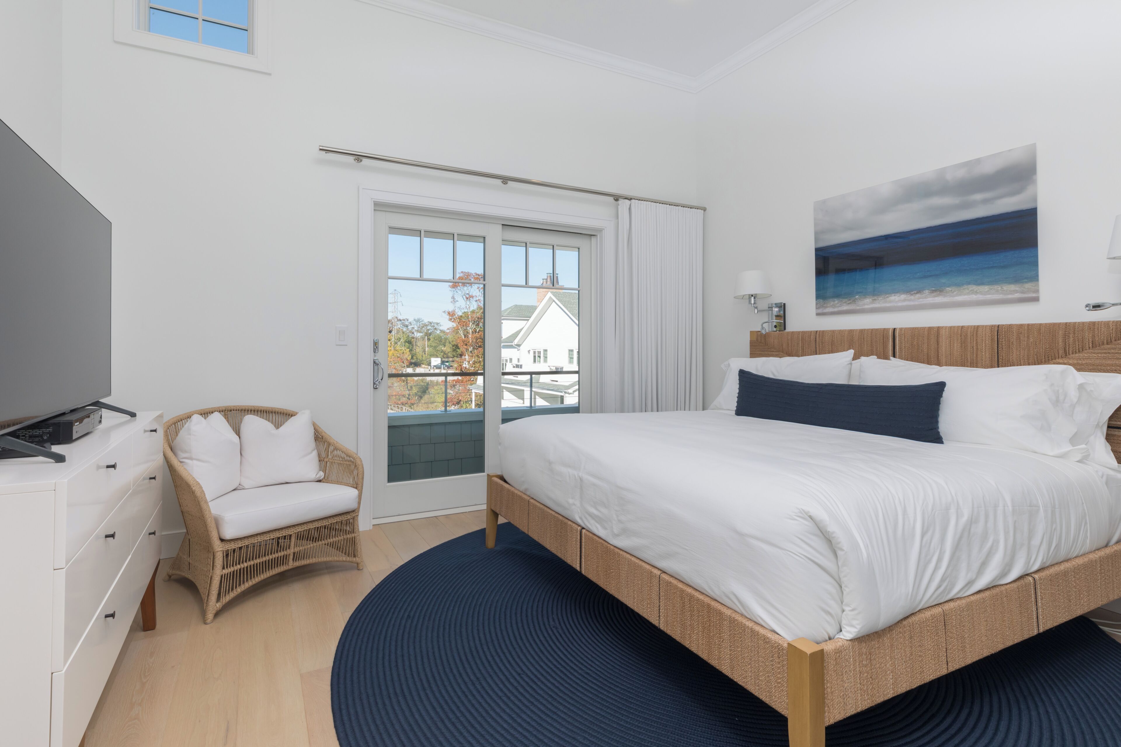 A bedroom with a bed, a chair, a television, a painting, and a view through a window.