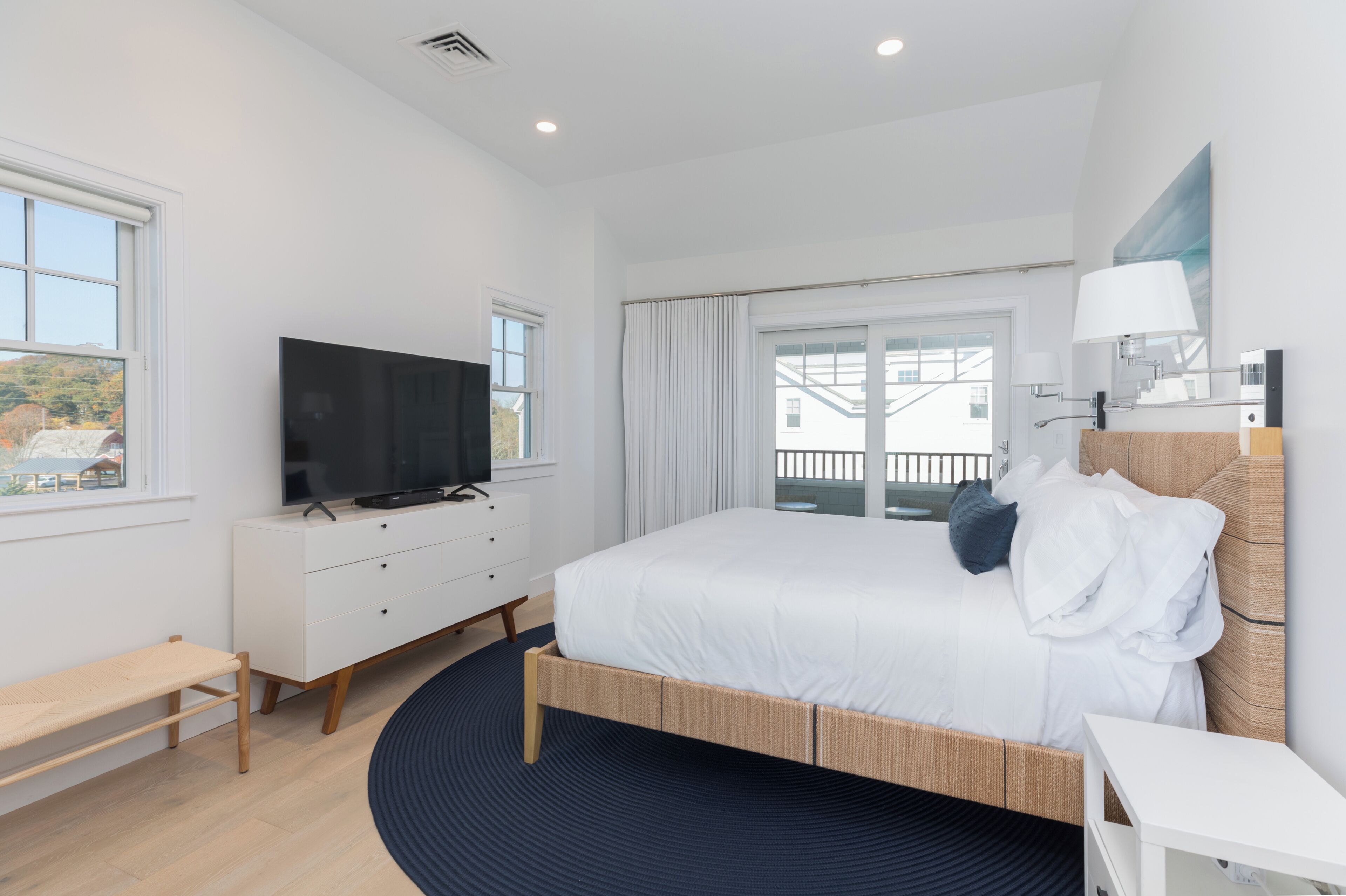 A modern hotel room with a large bed, a television on a white dresser, a window with a view, and a balcony.