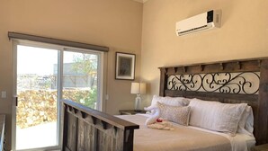 Un dormitorio con estructura de cama de madera, ropa de cama blanca, una mesita de noche con lámpara y vistas a través de un ventanal corredero.