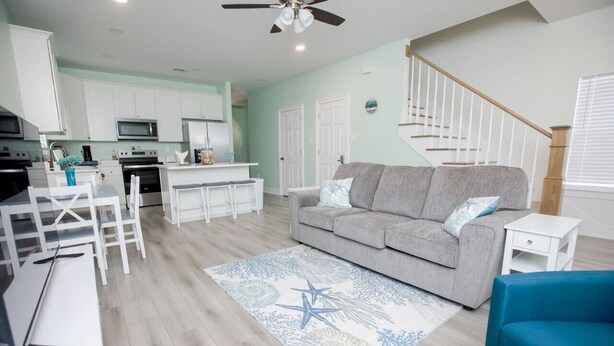 Una sala moderna con un sofá gris, una mesa de comedor con sillas, una zona de cocina con armarios blancos y una escalera.