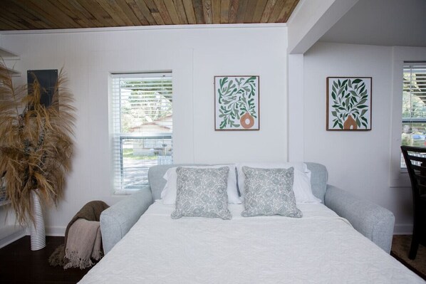 A bedroom with a bed, two pillows, a bedside table, a chair, and a window with blinds. - Mermaid Manor in Ocean Springs (Ocean Springs)