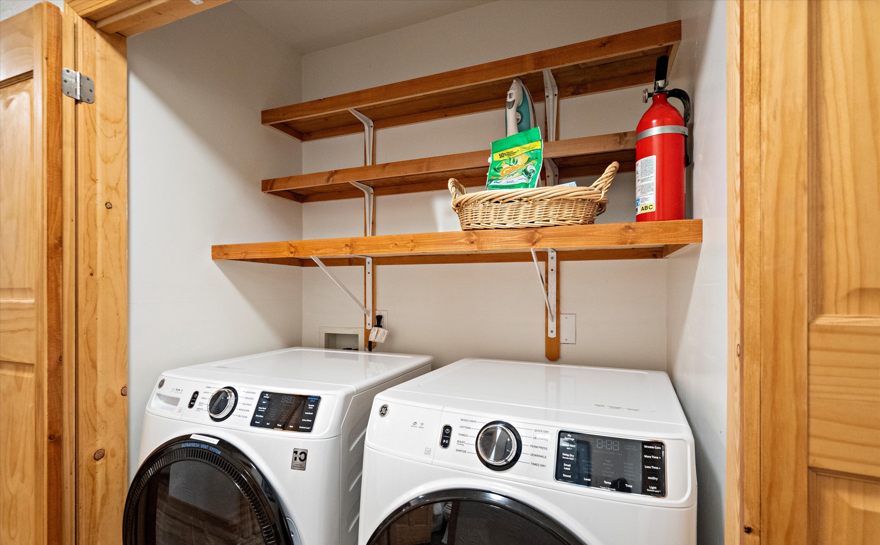 A laundry area with two front-loading washing machines, a fire extinguisher, and a shelf with a basket and a box.