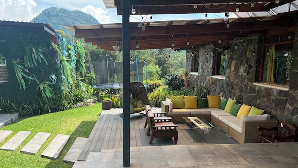 Un patio exterior con suelo de piedra, una pérgola de madera y una zona de estar con cojines.