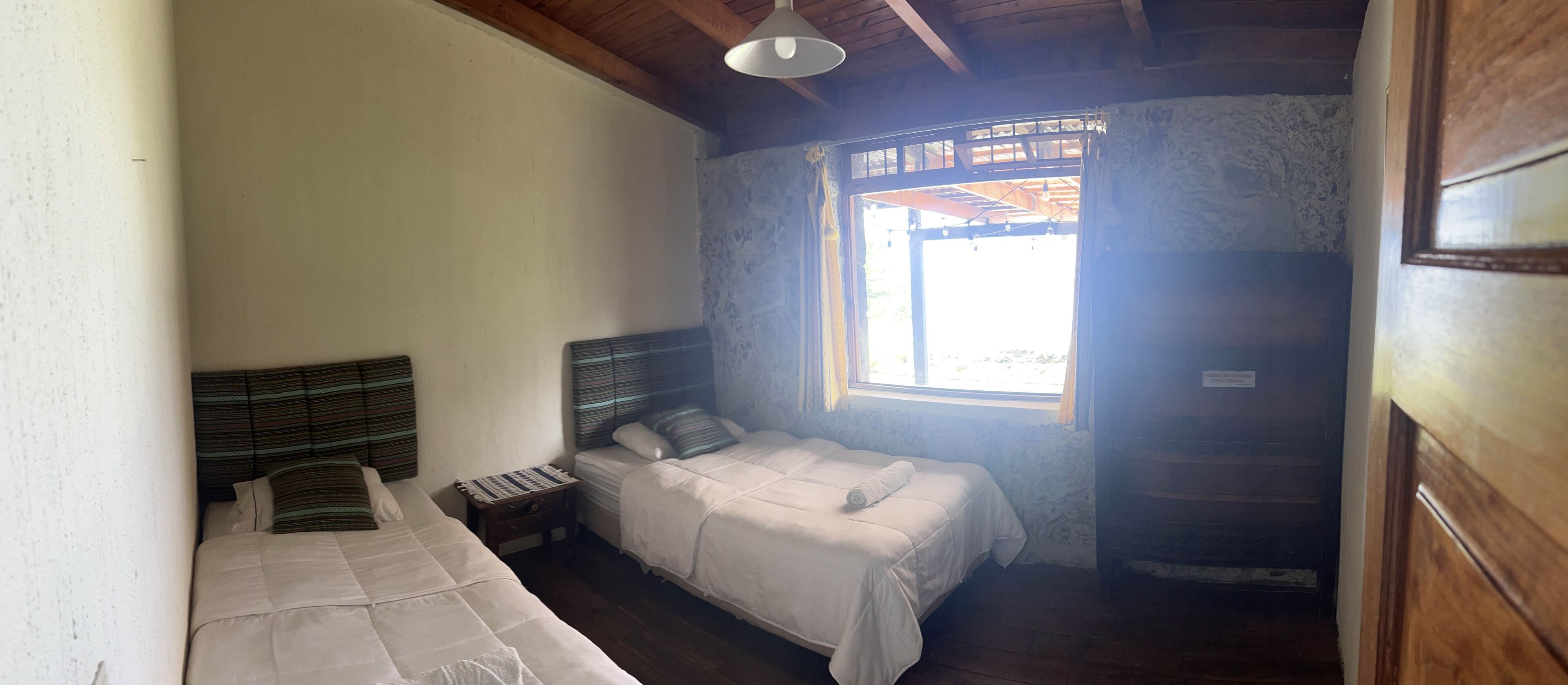 A small, simple room with two beds, a wooden ceiling, and a window with curtains.