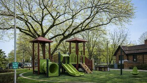 A playground with slides and climbing structures, surrounded by trees and a brick building. - The Oak Park Loft, Including Garage (Oak Park)
