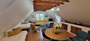 A cozy loft-style room with a bed, sofa, and dining area, featuring a wooden ceiling and a stained glass light fixture.