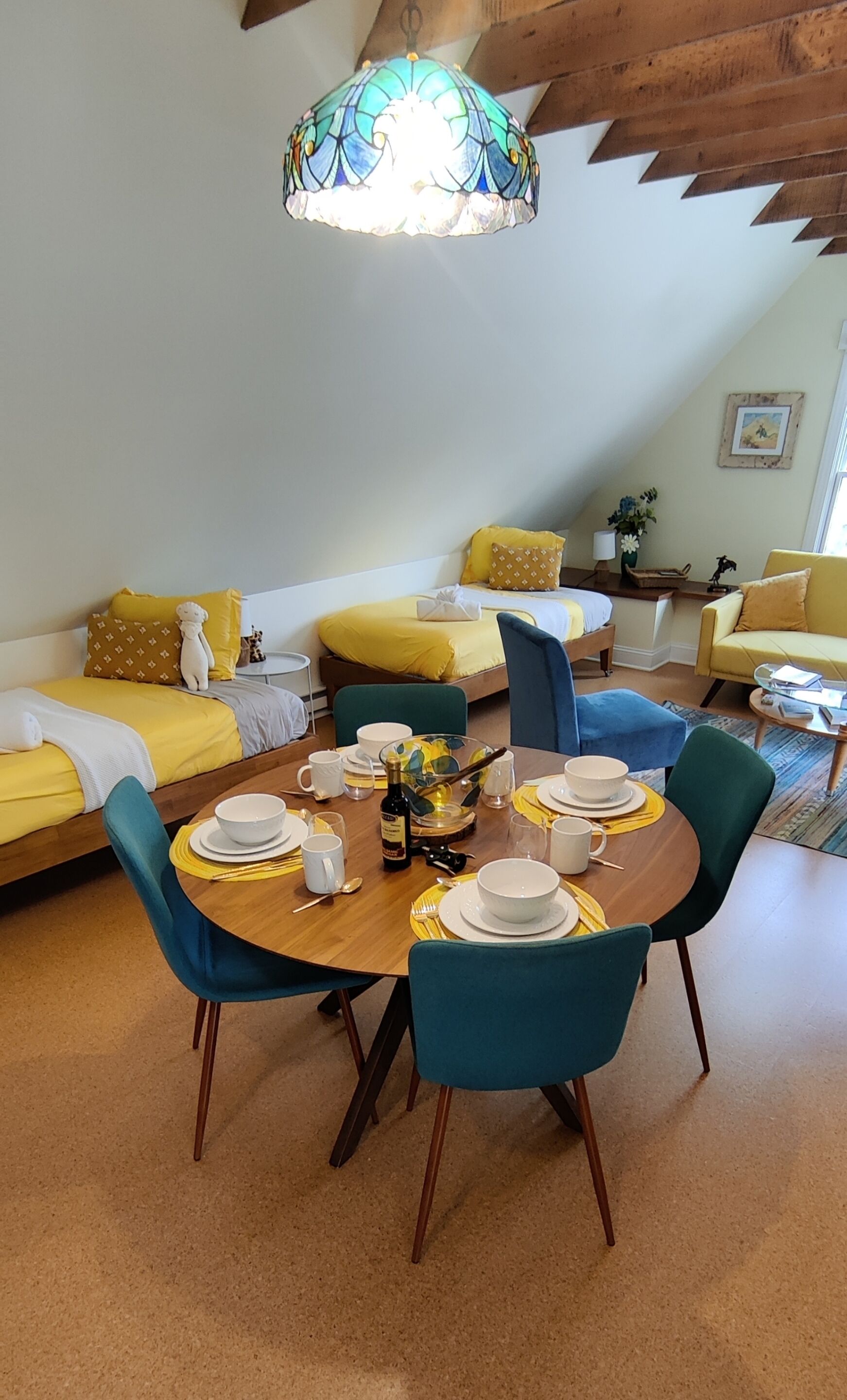 A dining area with a round table set for four, surrounded by teal chairs, in a room with yellow and blue furnishings.