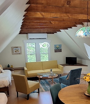 A cozy loft-style living room with a high ceiling, a yellow sofa, a glass coffee table, and a dining table with a vase of flowers.
