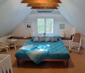 A bedroom with a sloped ceiling, a bed with a blue quilt, a window with a view of trees, and a chair by the window.