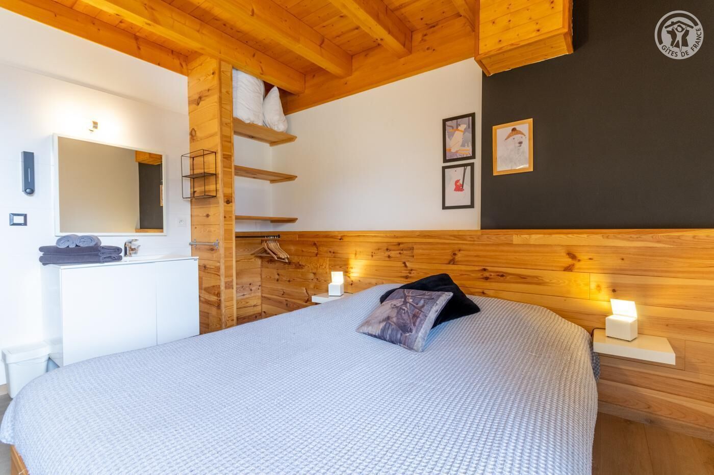 Une chambre avec une tête de lit en bois, un lit recouvert d’une courtepointe blanche à texture, une table de chevet avec une lampe, une étagère murale avec des serviettes et un miroir au-dessus du lavabo.