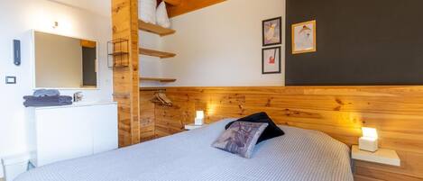 Une chambre avec une tête de lit en bois, un lit recouvert d’une courtepointe blanche à texture, une table de chevet avec une lampe, une étagère murale avec des serviettes et un miroir au-dessus du lavabo.