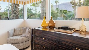 Habitación de esquina con un mueble de madera, un sillón estampado y un ventanal con vistas a palmeras y montañas.
