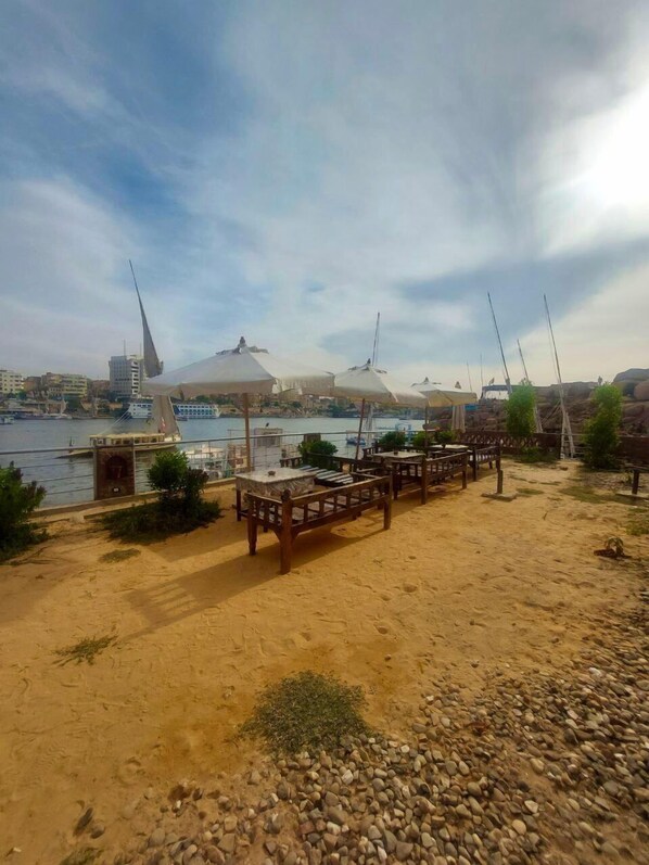 A beachside area with wooden benches and umbrellas, a sandy foreground, and a view of the sea and buildings in the background. - ANA-KA (Aswan)