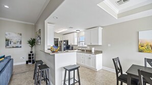 Une cuisine moderne avec des meubles blancs, un îlot central doté de tabourets, un coin repas avec une table et des chaises, et un espace de vie avec un canapé et une plante.