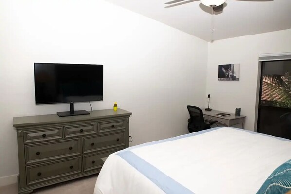 Un dormitorio con cama, televisor sobre un tocador, escritorio con silla y ventana con vista a un edificio.