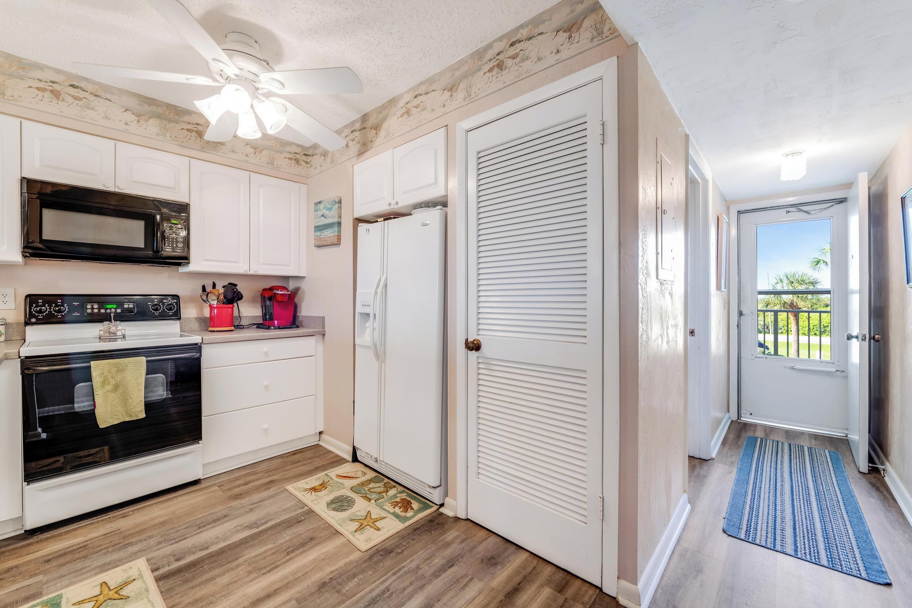 A compact kitchen with white cabinets, a microwave, a stove, and a refrigerator. There is a ceiling fan, a door leading outside, and a blue rug on the floor.