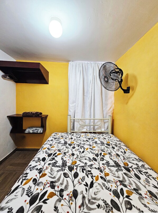 A small, brightly colored room with a bed, a wall-mounted fan, and a shelf with a pillow. - Casa Flores (Tehuacán)