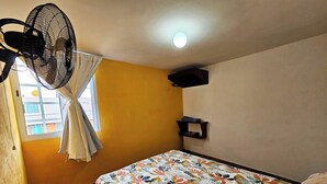 A room with a bed, a standing fan, a window with curtains, and a shelf with items. - Casa Flores (Tehuacán)