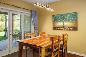 Un coin repas avec une table et des chaises en bois, un tableau représentant un arbre accroché au mur et une porte-fenêtre coulissante donnant sur un espace extérieur.