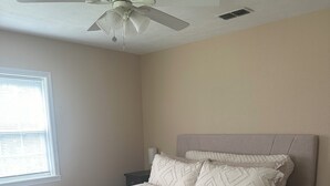 Une chambre à coucher avec un lit, un pouf gris, un ventilateur de plafond, une fenêtre et une table de chevet.