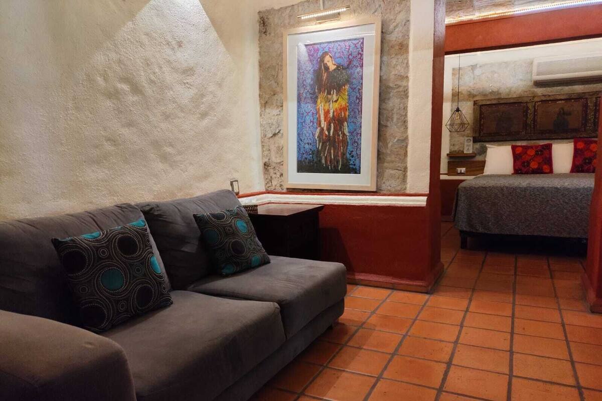 Un salon avec un canapé gris, des coussins à motifs, un tableau au mur et, au fond, un lit recouvert d’une couverture grise.