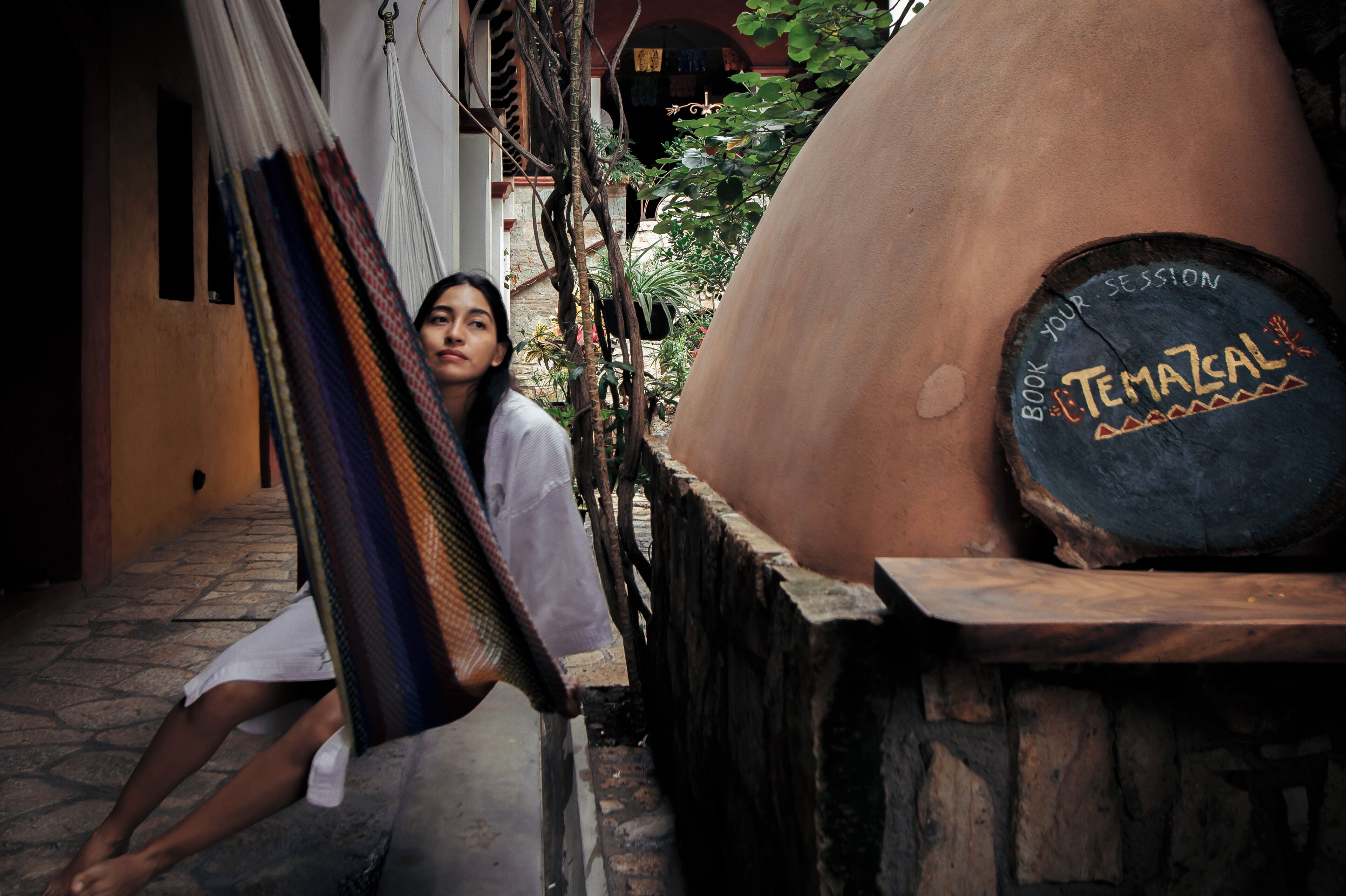 A person sitting on a hammock in a narrow alleyway with a large clay oven in the background.