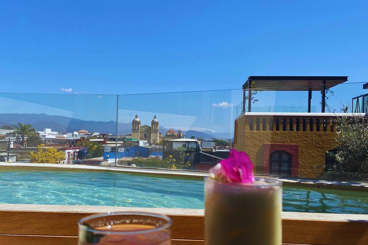 Vue d’un espace aménagé autour d’une piscine, avec deux boissons posées sur une table en bois, donnant sur un paysage urbain avec des bâtiments et des montagnes en arrière-plan.