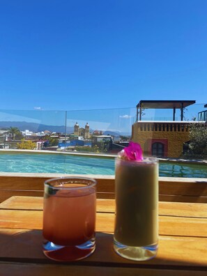 Vue d’un espace aménagé autour d’une piscine, avec deux boissons posées sur une table en bois, donnant sur un paysage urbain avec des bâtiments et des montagnes en arrière-plan.