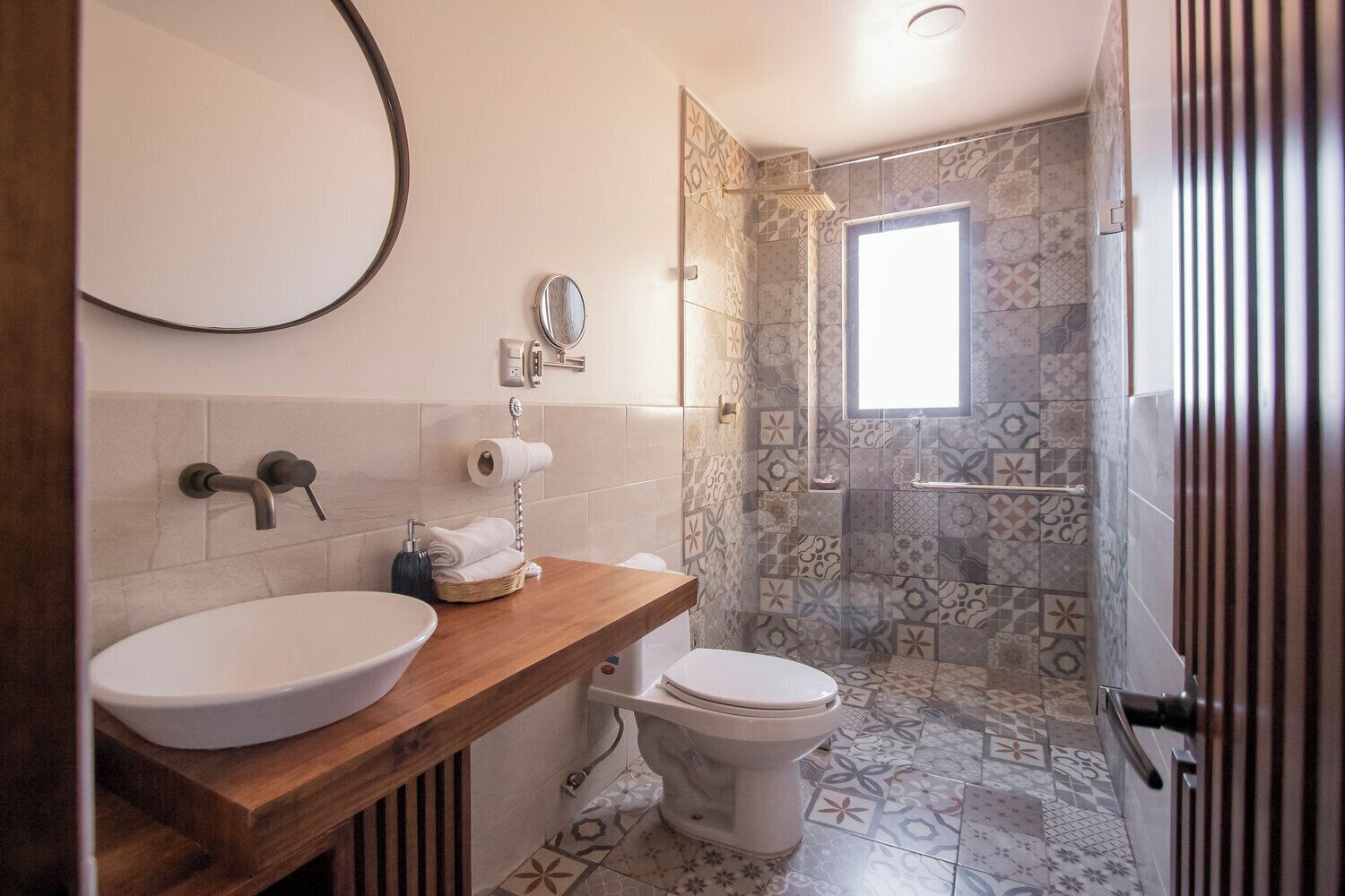 Un baño con tocador de madera, un lavabo blanco, un inodoro y una zona de ducha con azulejos estampados.