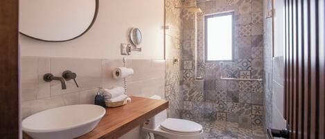 Un baño con tocador de madera, un lavabo blanco, un inodoro y una zona de ducha con azulejos estampados.