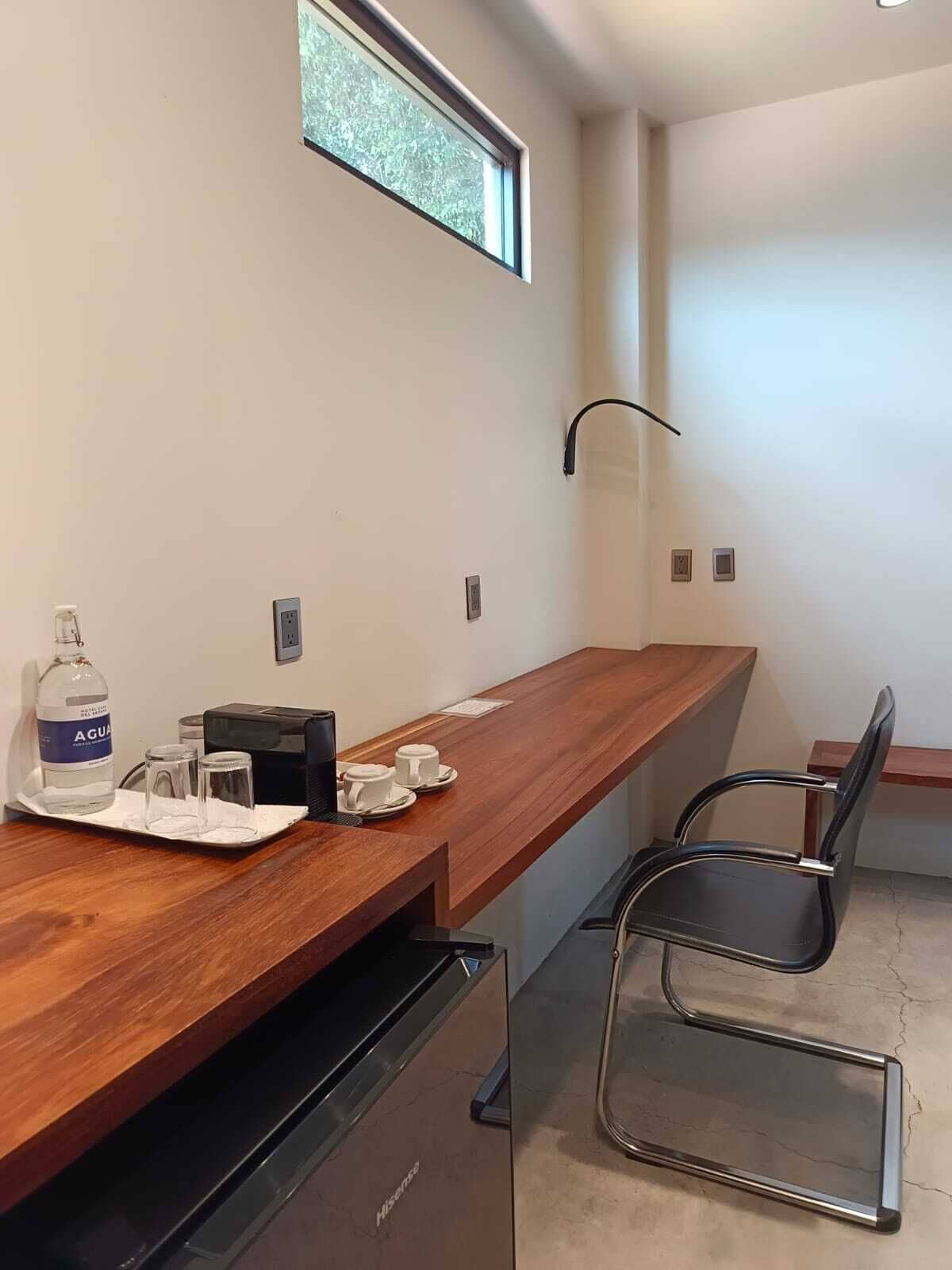 A modern hotel room with a wooden desk, a black chair, a minibar, and a window with a view of trees.