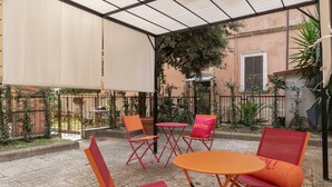 Une terrasse extérieure dotée de chaises et de tables pliantes, d’un espace abrité et offrant une vue sur un bâtiment avec une fenêtre et des plantes.