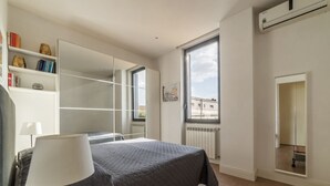 A bedroom with a bed, a bedside lamp, a large window, a radiator, and a bookshelf. - GuestHost - Modern and bright apartment of 60 sqm, able to accommodate up to 2 people, located on the second floor of a building without a lift (NOT suitable for disabled people).The property is located in an area well connected by public transport, just (Roma)