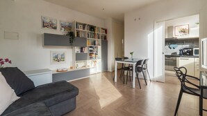 A modern living room with a sofa, a dining table with chairs, a bookshelf, and a kitchen area visible through an open door. - GuestHost - Modern and bright apartment of 60 sqm, able to accommodate up to 2 people, located on the second floor of a building without a lift (NOT suitable for disabled people).The property is located in an area well connected by public transport, just (Roma)