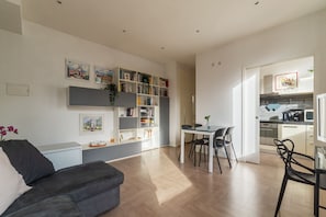 A modern living room with a sofa, a dining table with chairs, a bookshelf, and a kitchen area visible through an open door.