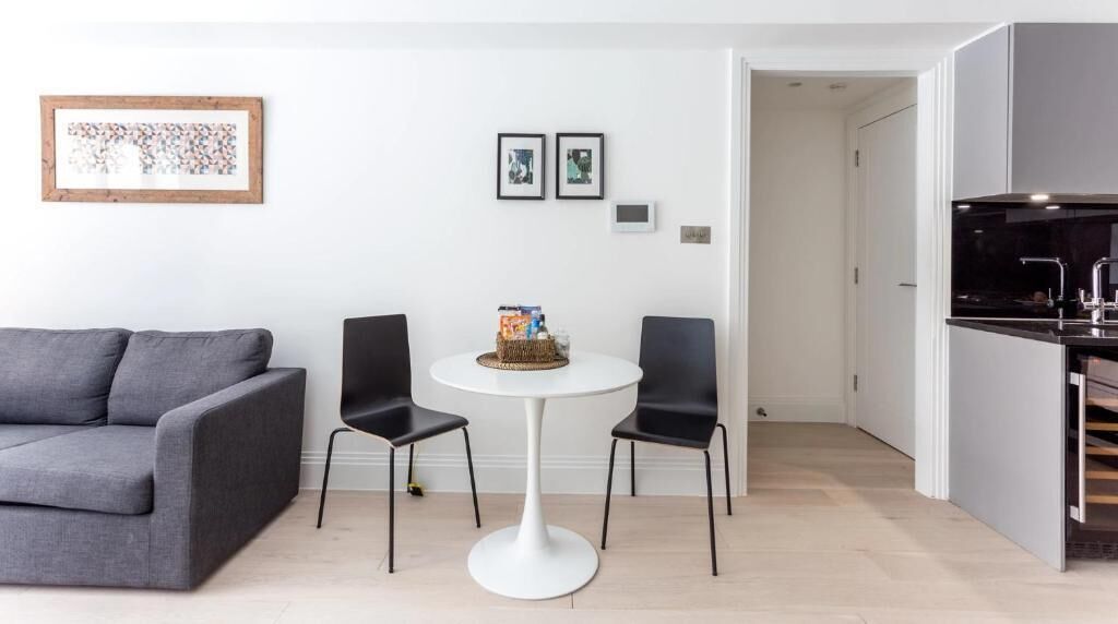 A modern living room with a grey sofa, a round white table with two black chairs, and a small bookshelf.