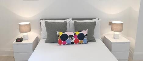 A neatly made bed with white bedding, two gray pillows, and a colorful pillow with a polka dot pattern. There are two bedside tables with lamps and a framed picture on the wall above the bed.