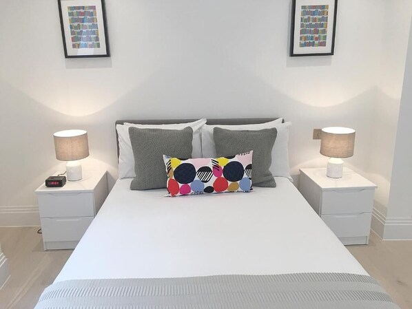 A neatly made bed with white bedding, two gray pillows, and a colorful pillow with a polka dot pattern. There are two bedside tables with lamps and a framed picture on the wall above the bed. - Oxford Circus Apartments (London)