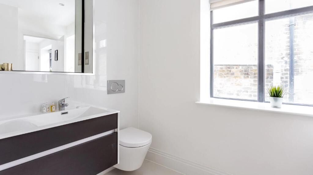 A modern bathroom with a white vanity, a toilet, and a window with a view of a brick building.
