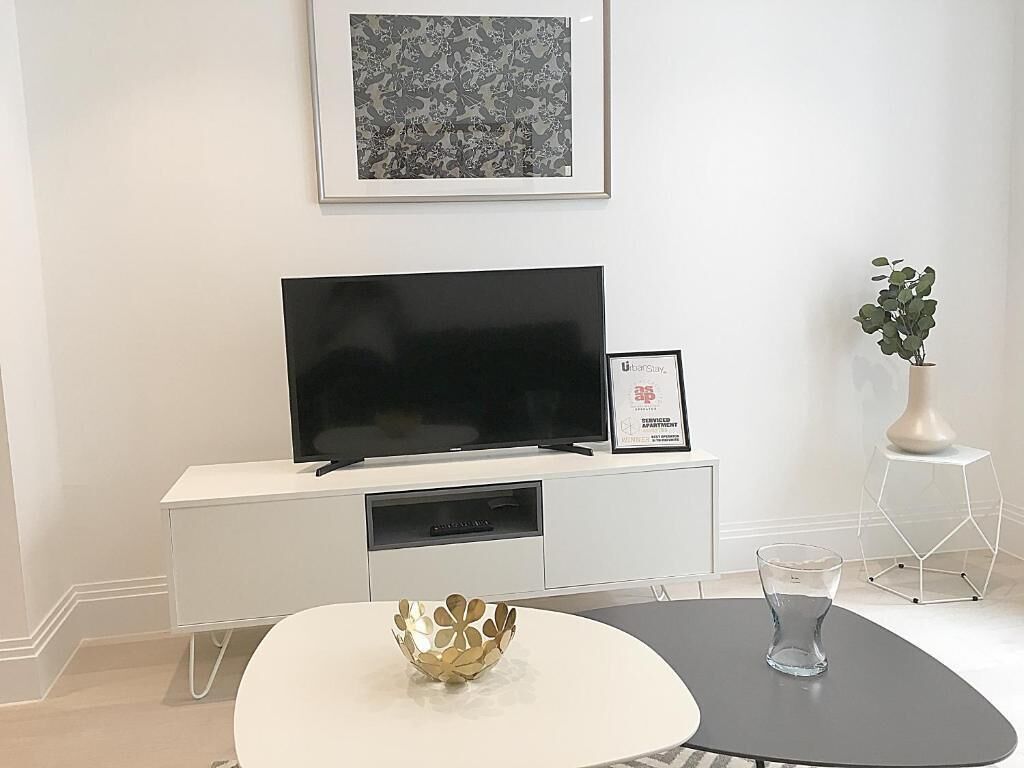 Una sala de estar moderna con una televisión de pantalla plana sobre un mueble blanco, un certificado enmarcado, un cuenco dorado decorativo, un florero de vidrio y una planta en maceta.
